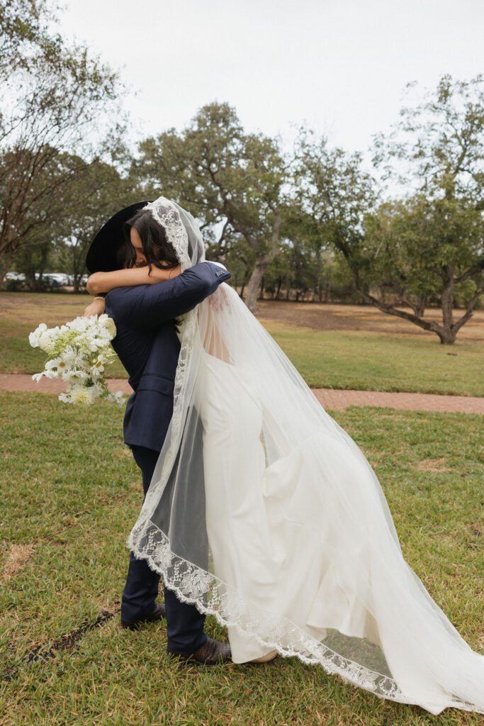 The groom, in his cowboy hat, embraces the bride in a romantic moment on the lawn, with her long lace veil draped gracefully around them.