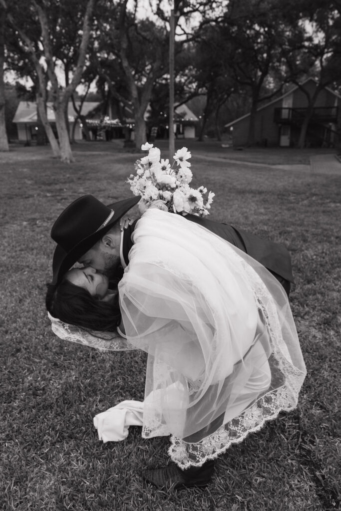 In a black-and-white shot, the groom dips the bride for a passionate kiss, with her veil flowing and the backdrop of oak trees adding depth.