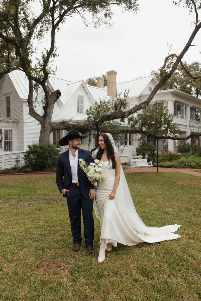 The bride and groom share a lighthearted moment in the garden, with the groom wearing a cowboy hat and the bride holding a bouquet of white flowers.